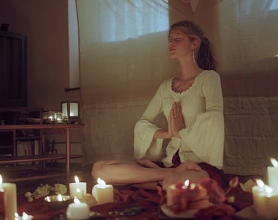 A young woman sitting cross-legged with  candles in a calm, dimly lit space.