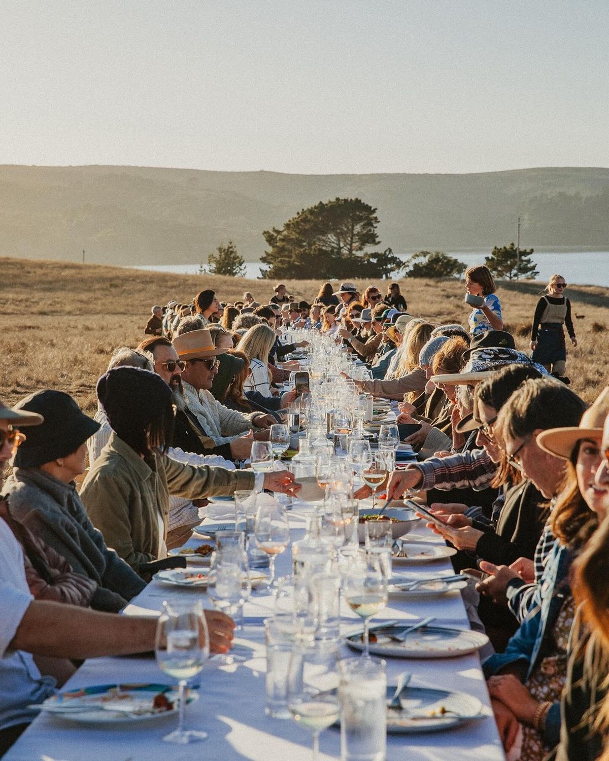 Eine lange Tafel unter freiem Himmel am Meer, an beiden Seiten sitzen Gäste beim Essen