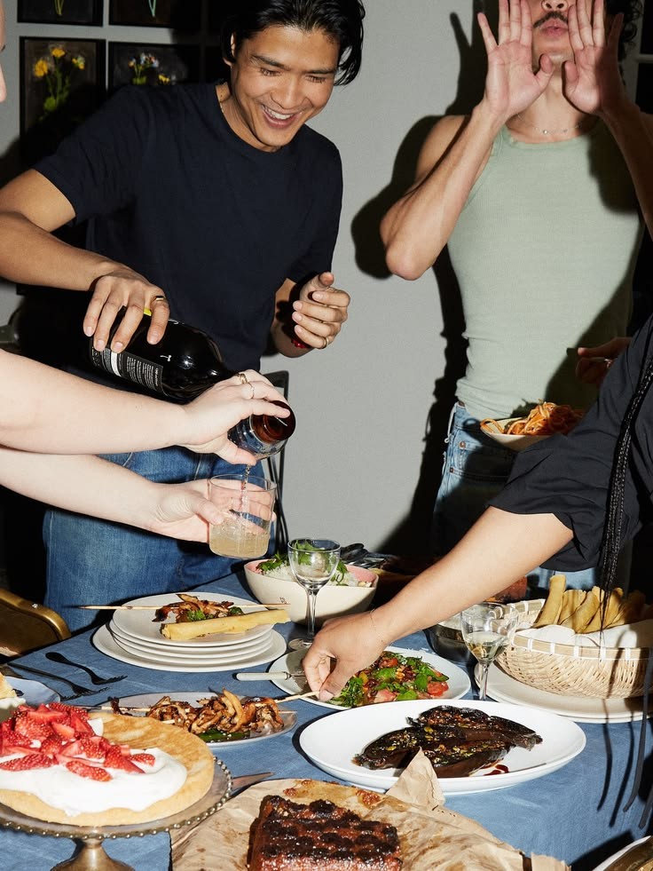 Freunde an einem Tisch voller Grillfleisch und Salate, einer schenkt Wein ein, alle lachen